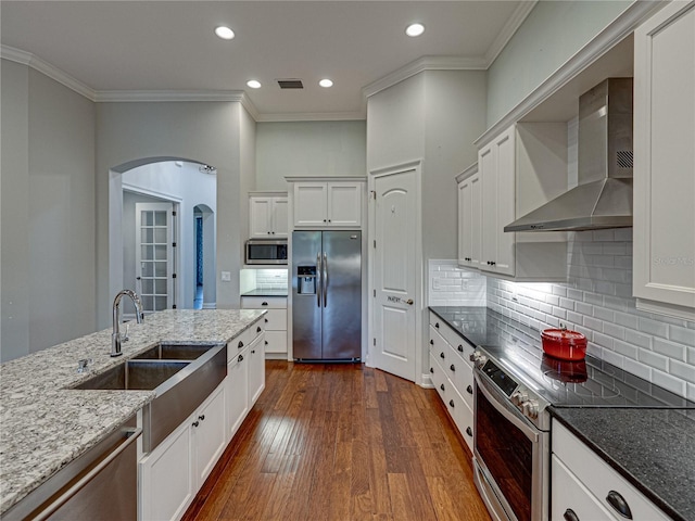 kitchen with dark wood-type flooring, dark stone counters, arched walkways, stainless steel appliances, and wall chimney exhaust hood