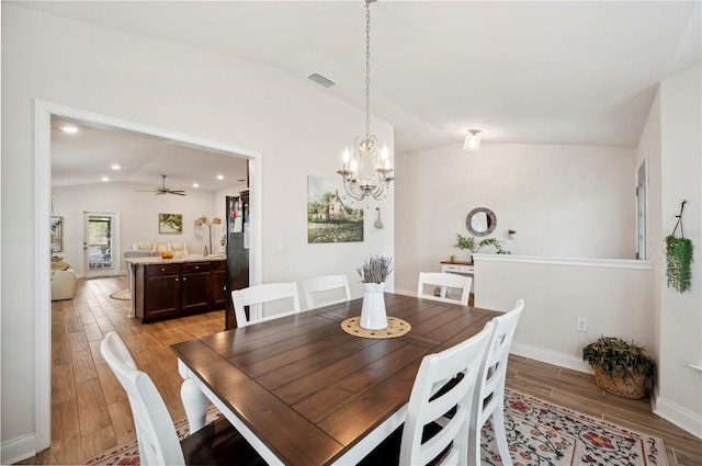 dining space featuring light wood-style flooring, ceiling fan with notable chandelier, visible vents, baseboards, and vaulted ceiling