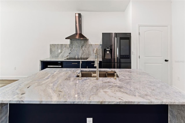 kitchen featuring a sink, backsplash, stainless steel fridge with ice dispenser, wall chimney range hood, and light stone countertops