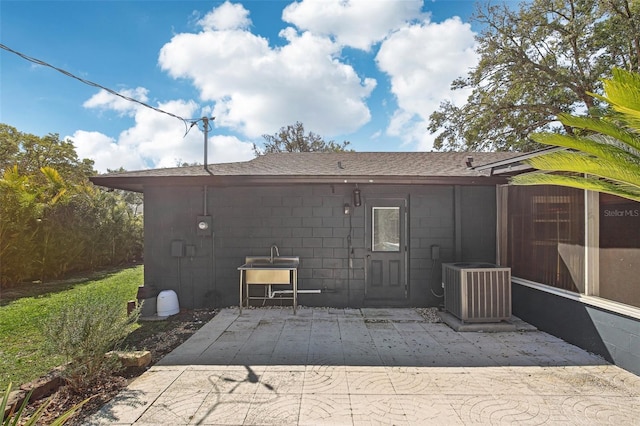 back of property with a shingled roof, cooling unit, concrete block siding, and a patio