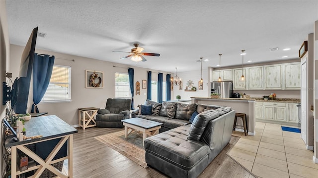 living room with light wood finished floors, visible vents, a textured ceiling, and ceiling fan with notable chandelier