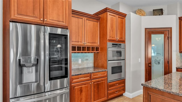 kitchen featuring baseboards, dark stone counters, appliances with stainless steel finishes, brown cabinets, and backsplash