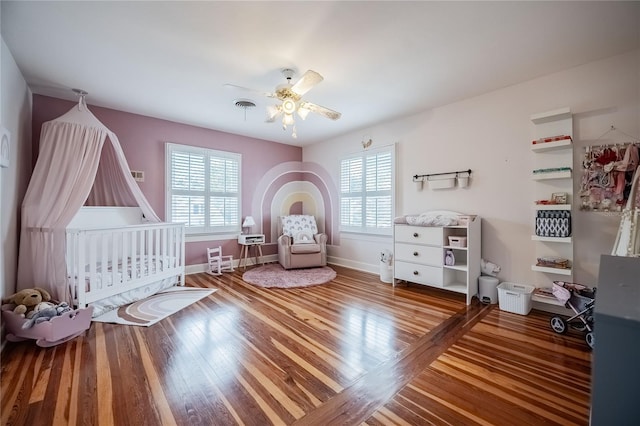 bedroom with visible vents, ceiling fan, baseboards, hardwood / wood-style floors, and a nursery area