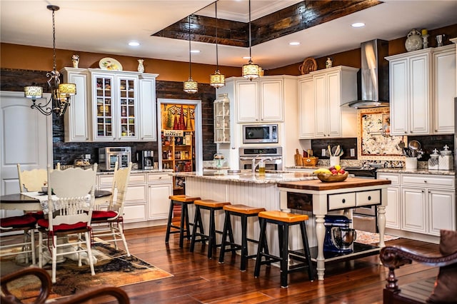 kitchen with a center island with sink, dark wood finished floors, stainless steel appliances, glass insert cabinets, and wall chimney range hood