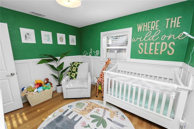 bedroom with a crib, wood finished floors, visible vents, and wainscoting