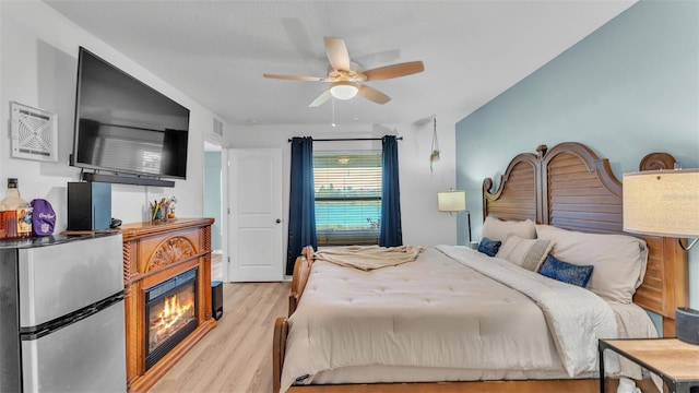 bedroom with a glass covered fireplace, light wood-style flooring, and a ceiling fan