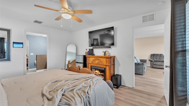 bedroom with visible vents, light wood-type flooring, a ceiling fan, and a glass covered fireplace