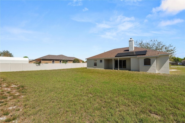 back of house featuring fence, roof mounted solar panels, a lawn, stucco siding, and a chimney