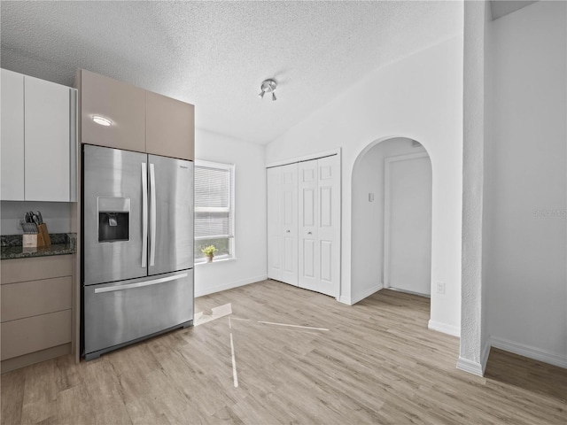 kitchen featuring stainless steel fridge with ice dispenser, vaulted ceiling, light wood-style flooring, arched walkways, and a textured ceiling