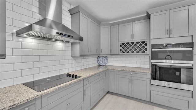 kitchen featuring wall chimney range hood, double oven, black electric cooktop, gray cabinetry, and tasteful backsplash