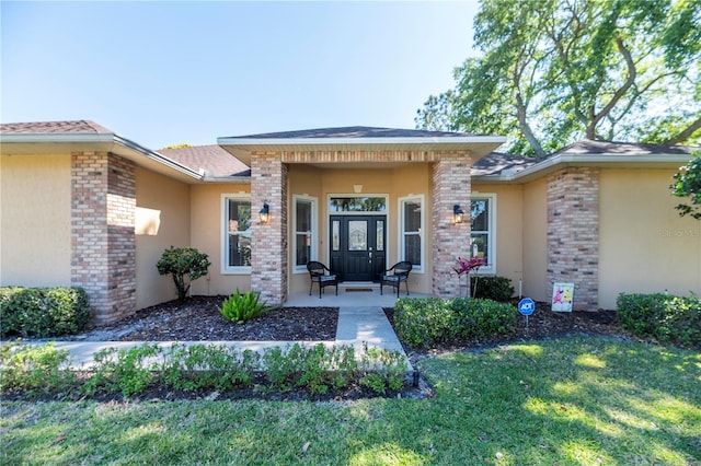 entrance to property featuring brick siding, a yard, roof with shingles, and stucco siding