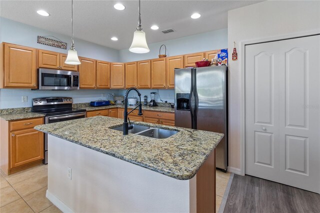 kitchen featuring hanging light fixtures, a sink, appliances with stainless steel finishes, a center island with sink, and light stone countertops