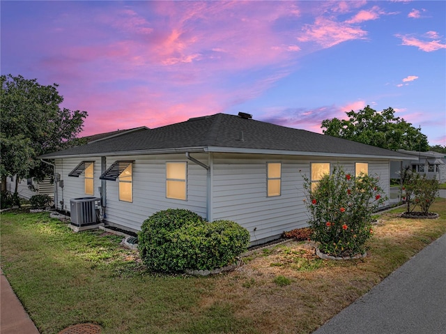 property exterior at dusk with central AC unit, a yard, and roof with shingles