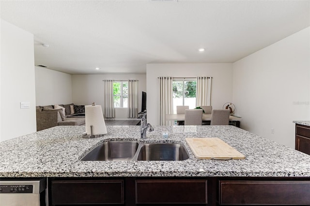 kitchen featuring stainless steel dishwasher, light stone counters, dark brown cabinets, a sink, and open floor plan