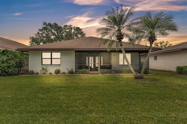 back of house at dusk with a lawn, a shingled roof, a sunroom, and stucco siding