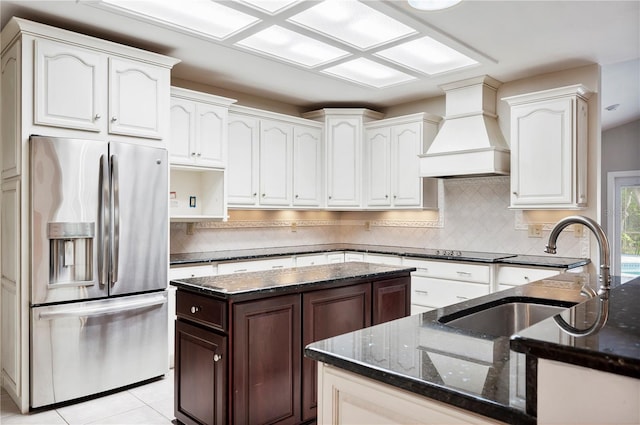 kitchen featuring stainless steel refrigerator with ice dispenser, premium range hood, a sink, and dark stone counters