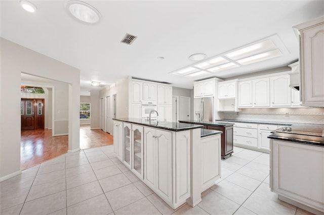 1104 Kells Ct, Lakeland, FL 33813 - kitchen with stainless steel fridge, a kitchen island with sink, light tile patterned floors, glass insert cabinets, and white cabinetry
