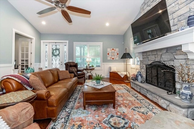 living area featuring ceiling fan, a stone fireplace, vaulted ceiling, wood finished floors, and a wainscoted wall
