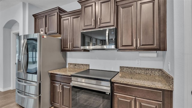 kitchen featuring stainless steel appliances, arched walkways, light stone countertops, and dark brown cabinets