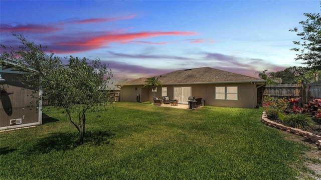 back of house at dusk with fence, stucco siding, an outbuilding, a lawn, and a patio
