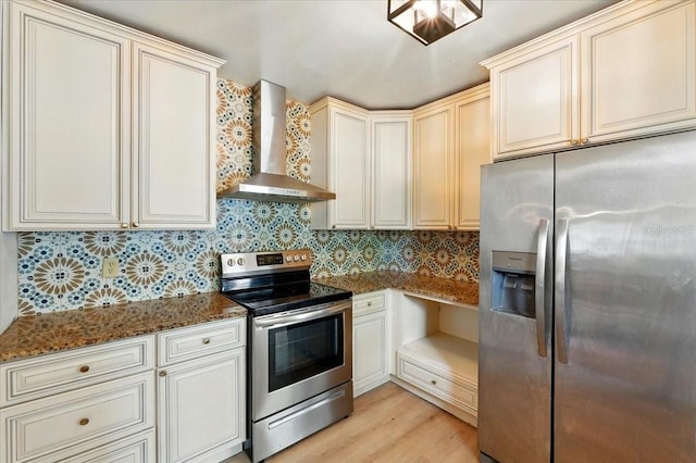 kitchen with wall chimney range hood, dark stone counters, light wood finished floors, decorative backsplash, and appliances with stainless steel finishes