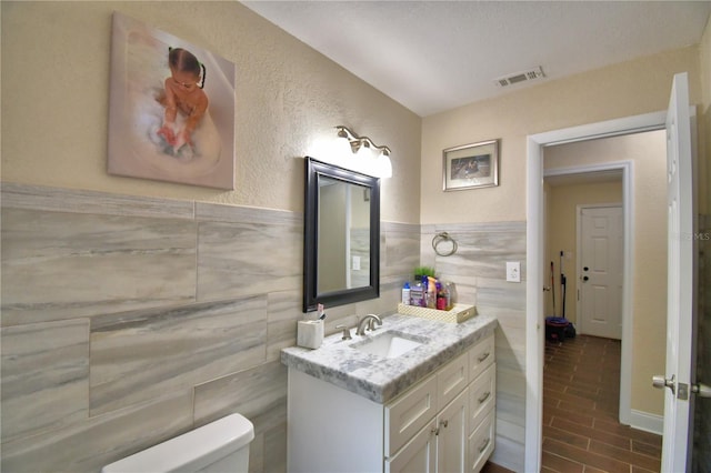bathroom with vanity, toilet, a wainscoted wall, tile walls, and visible vents