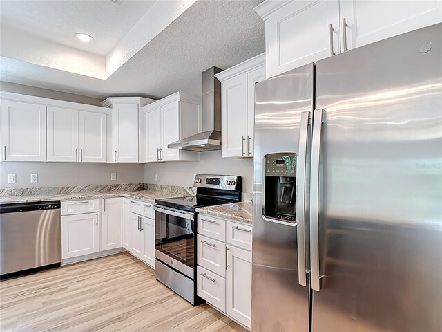 kitchen featuring stainless steel appliances, wall chimney exhaust hood, white cabinetry, light stone countertops, and light wood finished floors
