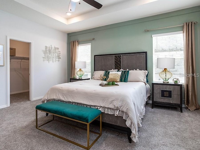 carpeted bedroom featuring a closet, a ceiling fan, a tray ceiling, a walk in closet, and baseboards