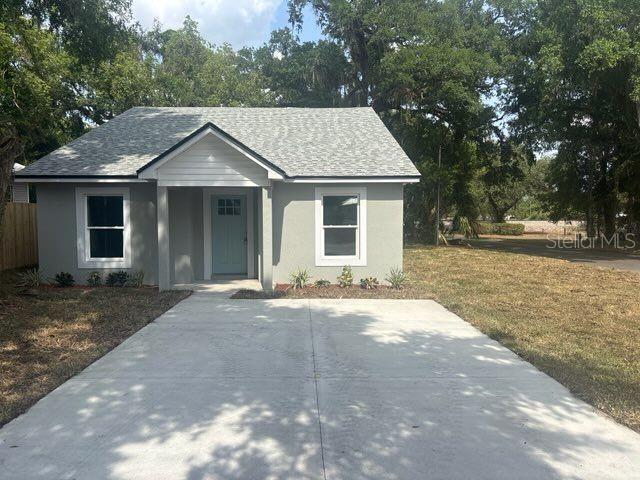 ranch-style house with a front lawn, roof with shingles, stucco siding, and view of wooded area