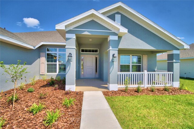 view of exterior entry featuring a porch, stucco siding, a yard, and a shingled roof