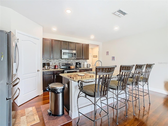 kitchen with appliances with stainless steel finishes, a kitchen bar, dark brown cabinets, dark wood-style flooring, and light stone countertops