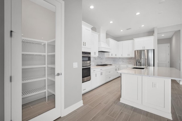 kitchen featuring white cabinetry, stainless steel appliances, light hardwood / wood-style flooring, premium range hood, and a kitchen island with sink