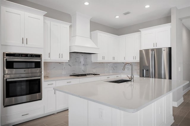 kitchen with backsplash, custom range hood, a kitchen island with sink, and appliances with stainless steel finishes