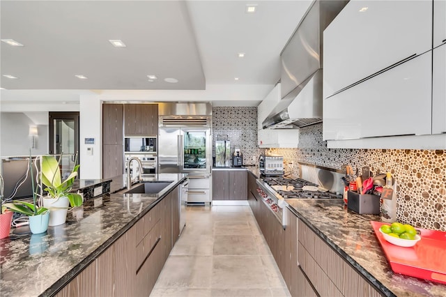 kitchen featuring wall chimney exhaust hood, sink, built in appliances, dark stone counters, and decorative backsplash