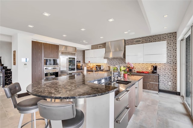 kitchen featuring a large island, wall chimney range hood, sink, white cabinetry, and built in appliances