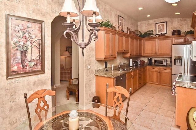 kitchen with sink, dark stone countertops, appliances with stainless steel finishes, light tile flooring, and a notable chandelier