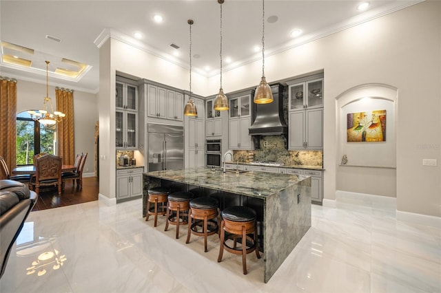 kitchen with appliances with stainless steel finishes, dark stone counters, gray cabinetry, custom range hood, and a kitchen island with sink