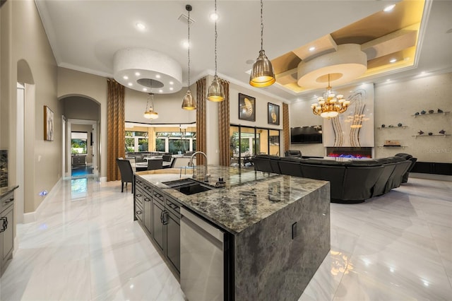 kitchen featuring dark stone counters, a raised ceiling, crown molding, an island with sink, and decorative light fixtures