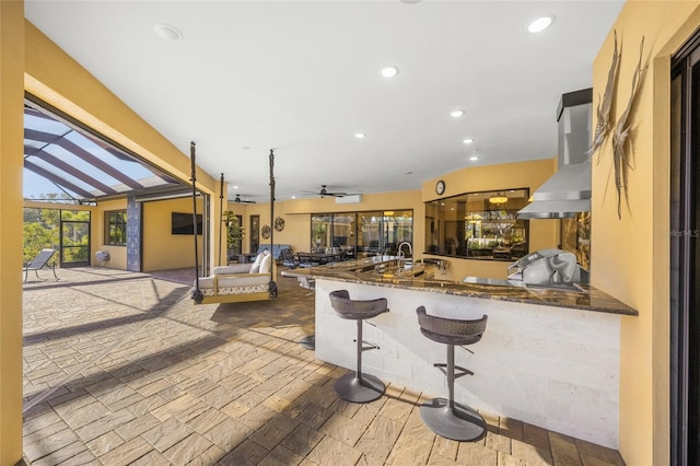 kitchen with ceiling fan, kitchen peninsula, dark stone countertops, a kitchen bar, and island range hood