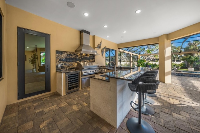 kitchen featuring a kitchen bar, sink, wall chimney range hood, dark stone countertops, and wine cooler