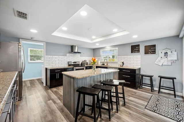 kitchen with wall chimney exhaust hood, stainless steel appliances, a center island, and light hardwood / wood-style floors