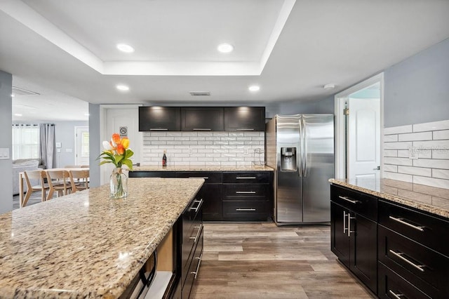 kitchen with stainless steel refrigerator with ice dispenser, light stone counters, light hardwood / wood-style floors, tasteful backsplash, and a raised ceiling