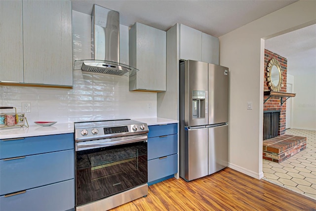 kitchen with appliances with stainless steel finishes, light wood-type flooring, blue cabinets, wall chimney exhaust hood, and tasteful backsplash