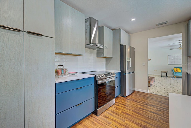 kitchen with appliances with stainless steel finishes, light hardwood / wood-style flooring, wall chimney range hood, ceiling fan, and blue cabinets