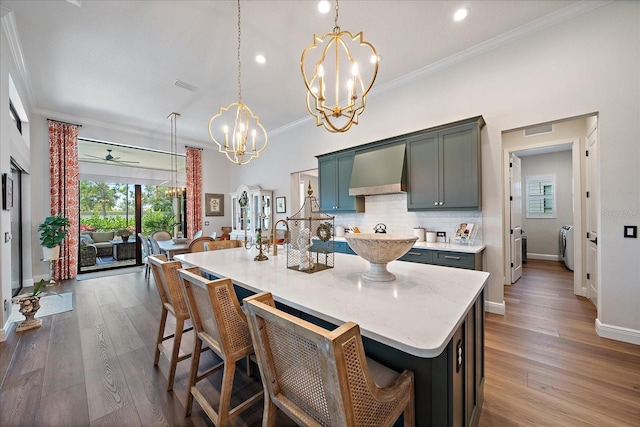 kitchen featuring ornamental molding, an island with sink, pendant lighting, washing machine and dryer, and wall chimney range hood