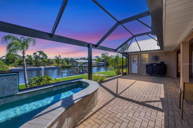 pool at dusk with glass enclosure, a patio area, a dock, and a water view