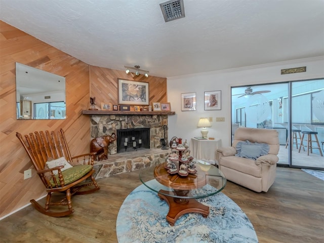 living room with ceiling fan, a stone fireplace, wood walls, hardwood / wood-style floors, and ornamental molding