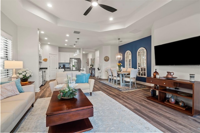 living room featuring ceiling fan with notable chandelier, a raised ceiling, and dark wood-type flooring