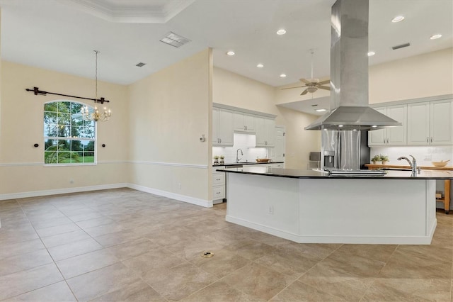 kitchen with dark countertops, ceiling fan with notable chandelier, island exhaust hood, white cabinets, and a sink