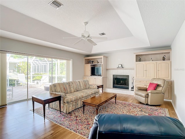 living room featuring a tile fireplace, ceiling fan, light hardwood / wood-style floors, a textured ceiling, and a raised ceiling